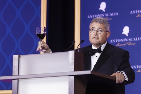 Leonard Leo, standing at a podium, holds up his wine glass and toasts a group of people. 
