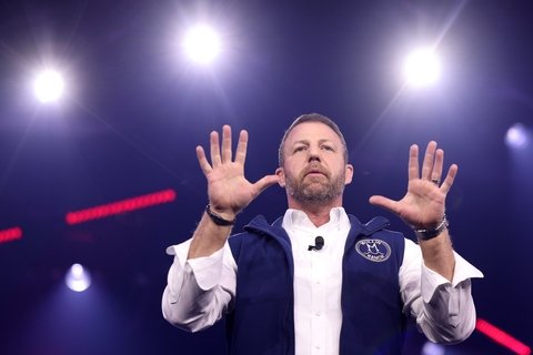 man in a white shirt and blue vest stands with hands raised with spotlights in the background