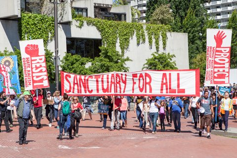 protestors march with a white banner with red text reading "sanctuary for all"