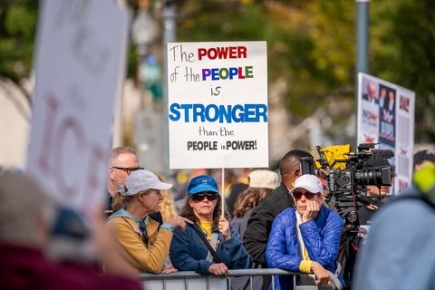 a group of people behind a barrier. The person in the center holds a sign that reads "the power of the people is stronger than the people in power."