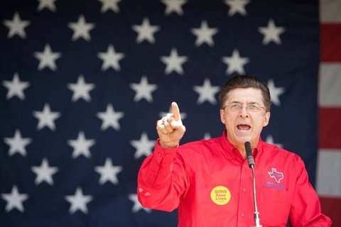 A man in a red jacket speaking into a mic with an American flag behind him.