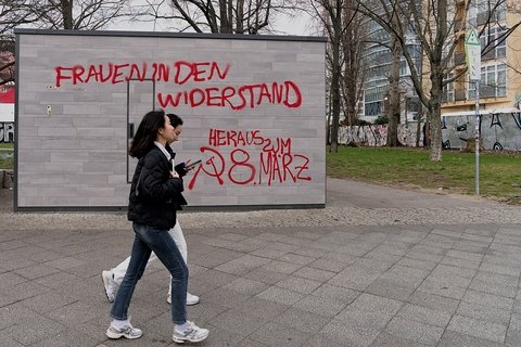 Two femme-bodied people walking through a park in front of some graffiti. Berlin-Friedrichshain, Warschauer Straße. Toilettenhäuschen auf dem Parkgelände Warschauer Straße Ecke Mühlenstraße, mit aufgesprühter, roter Aufschrift „Frauen in den Wiederstand heraus zum 8. März“. (Berlin-Friedrichshain, Warschauer Straße. Toilet block in the park at the corner of Warschauer Straße and Mühlenstraße, with the spray-painted red inscription "Women in the resistance, out on March 8th.")