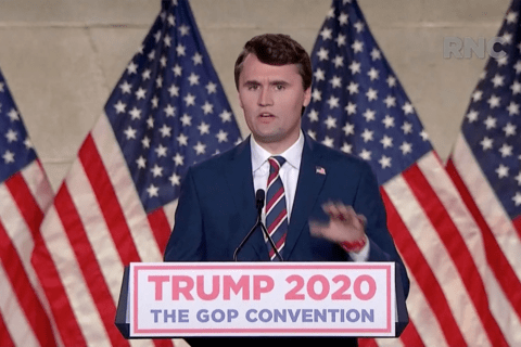 A man in a suit standing in front of American flags, speaking into a mic