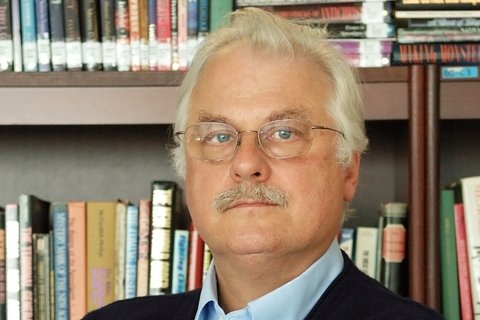 a man with white hair, glasses, and a moustache stands in front of a book case