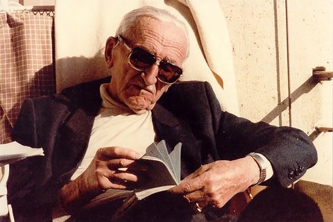 old man with sunglasses and white hair reads a book in the sun