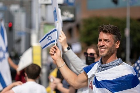 white man waves israeli flag while wearing another israeli flag as a cape