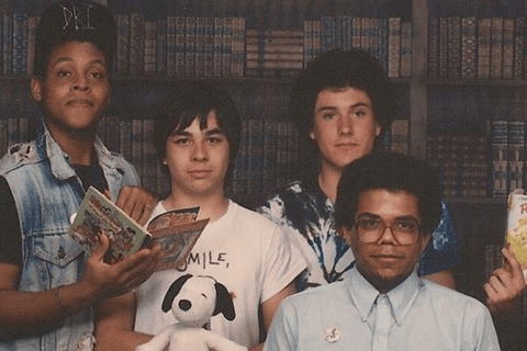 Four young people of various skin colors standing in front of long bookshelf, some holding books.