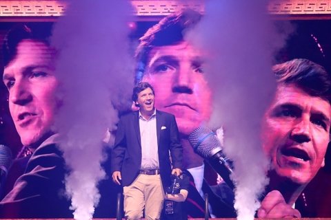 Tucker Carlson walks onto stage with giant images of his head behind him. 