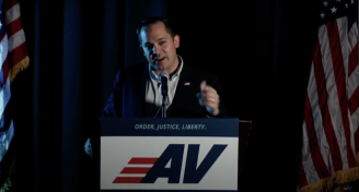 A man in blue suit standing at a podium with the letters AV on it speaking into a mic with two American flags behind him.