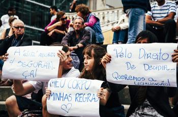People sitting on stairs. Three people hold signs in Portugese, written with blue ink on white paper.