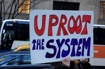 A person holds up a sign that reads "Uproot the system"