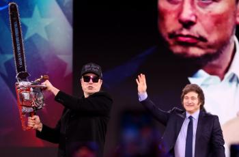 Two men standing in front of projector image. One man is waving at the crowd, the other man is holding a chainsaw