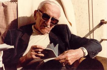 old man with sunglasses and white hair reads a book in the sun