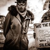 Two men in winter clothes, one holding a sign that reads "We the people will not be silenced"