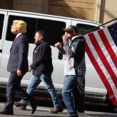 Three men walking; one wears a Trump mask and one carries an American flag