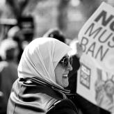A woman in a hijab smiling, holding a poster that says No Muslim Ban