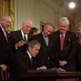 Presdient George Bush signing something, with men standing around him.