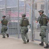 Three men in army training suits running along a large metal fence