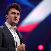 A young white man, Charlie kirk, in a suit, holding a mic.