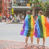 Two people with rainbow flags on their backs, looking into a street.