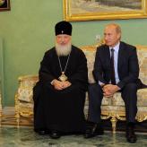 A man in a black robe and a black hate with a cross on it sitting next to Vladimir Putin on a golden couch.