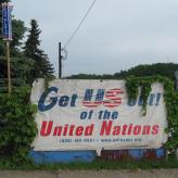 A sign covered by plants. The sign reads "Get US out! of the United Nations (888) JBS USA1"