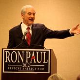 A man in a suit talking into a mic and pointing to his left. A sign on the podium says "Ron Paul, 2012, Restore America Now"