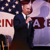 A bald man in a suit, standing in front of an American flag, talking into a mic.