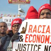 Woman in hat and gloves holds sign reading "Racial Economic Justice Now."