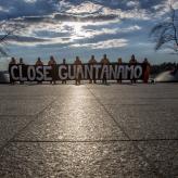 A row of people in orange jumpsuits holding a sign that says "close Guantanamo"