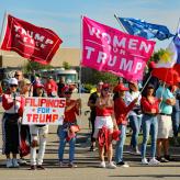 A group of women in red hats hold flags reading "Women for Trump," "Filipinos for Trump," and "Trump pence"