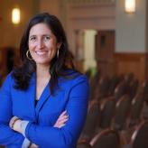 Smiling woman with long brown hair, gold hoop earrings, and a blue blazer, arms crossed