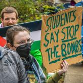 A person wearing a mask and glasses holds a sign reading "Students say stop the bombs"