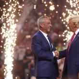 Two men shaking hands with fireworks behind them.