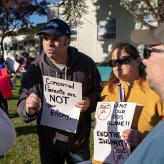 A man in a black sweatshirt and hat holding a sign that says  "Concerned Parents are NOT your enemy" and a woman in a yellow sweatshirt holding a sign that says "Leave our kids alone!! End the Insanity" with a stop sign over SOGI 123.