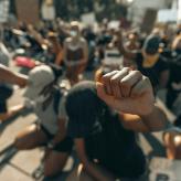 Focus on a raised fist, background of people kneeling in protest.