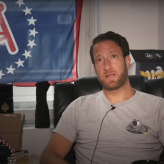 A man in a grey T-shirt sitting in a chair with a flag behind him with the Barstool logo