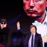 Two men standing in front of projector image. One man is waving at the crowd, the other man is holding a chainsaw