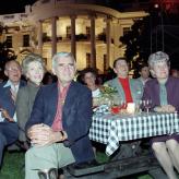 Six people sitting around a gingham table cloth on a picnic table with the white house in the background
