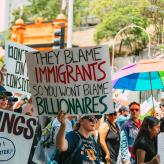 a person holding a sign that says "they blame the immigrants so you won't blame billionaires"