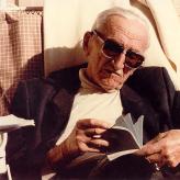 old man with sunglasses and white hair reads a book in the sun