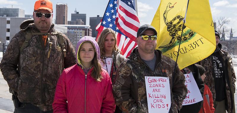 Small group poses for camera with US and Gadsden flags. In front, a child in pink jacket stands beside man wearing a brown camo jacket with sign pinned on it. Sign reads: "GUN FREE ZONES ARE KILLING OUR KIDS"