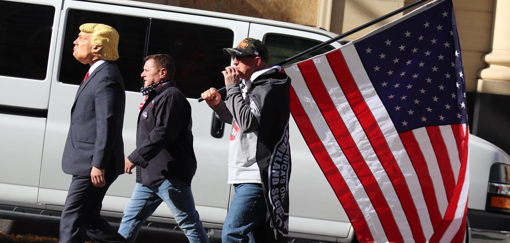 Three men walking; one wears a Trump mask and one carries an American flag