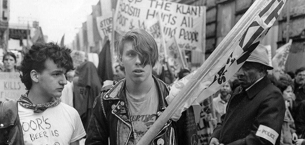 Two people at a protest. One is holding a flag, wearing a leather jacket. The other man is wearing a white t-shirt with anti-nazi writing. 