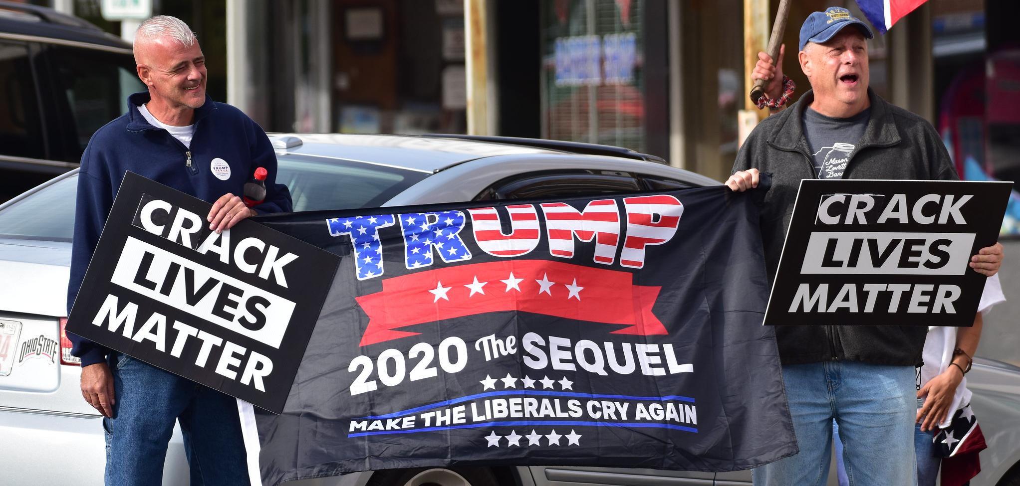 Two men holding a flag that says Trump 2020 The Sequel and banners saying "crack lives matter"