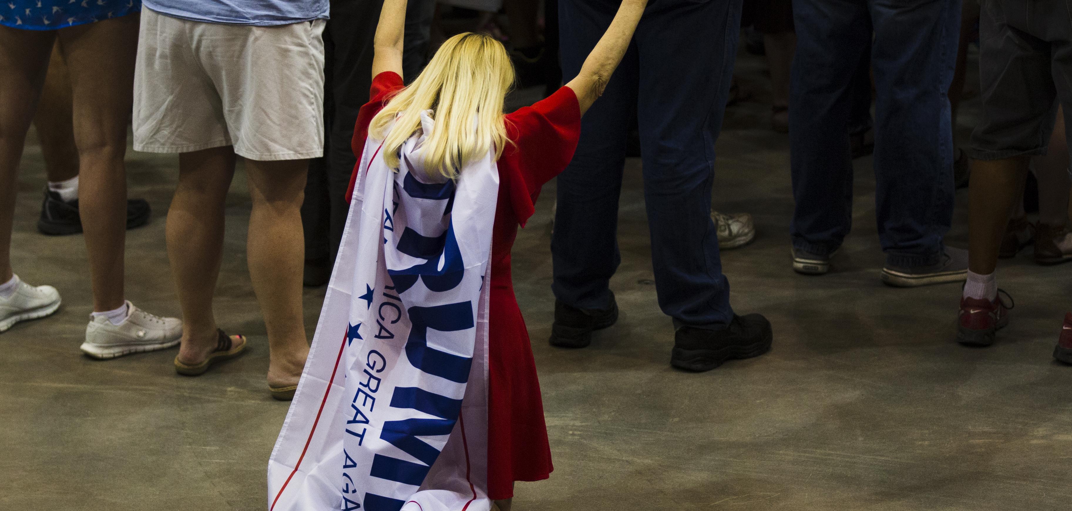 A white woman with blond hair, kneeling with her arms in the air, a Trump flag tied around her neck and hanging down her back