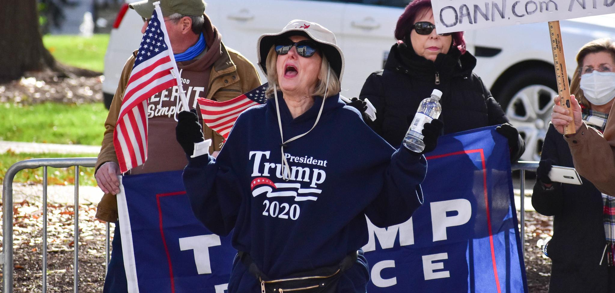 A White woman with blond hair, wearing a hat. She has a Trump 2020 sweatshirt on, and is holding an Amerian Flag and a water bottle. Her mouth is open like she's shouting something.
