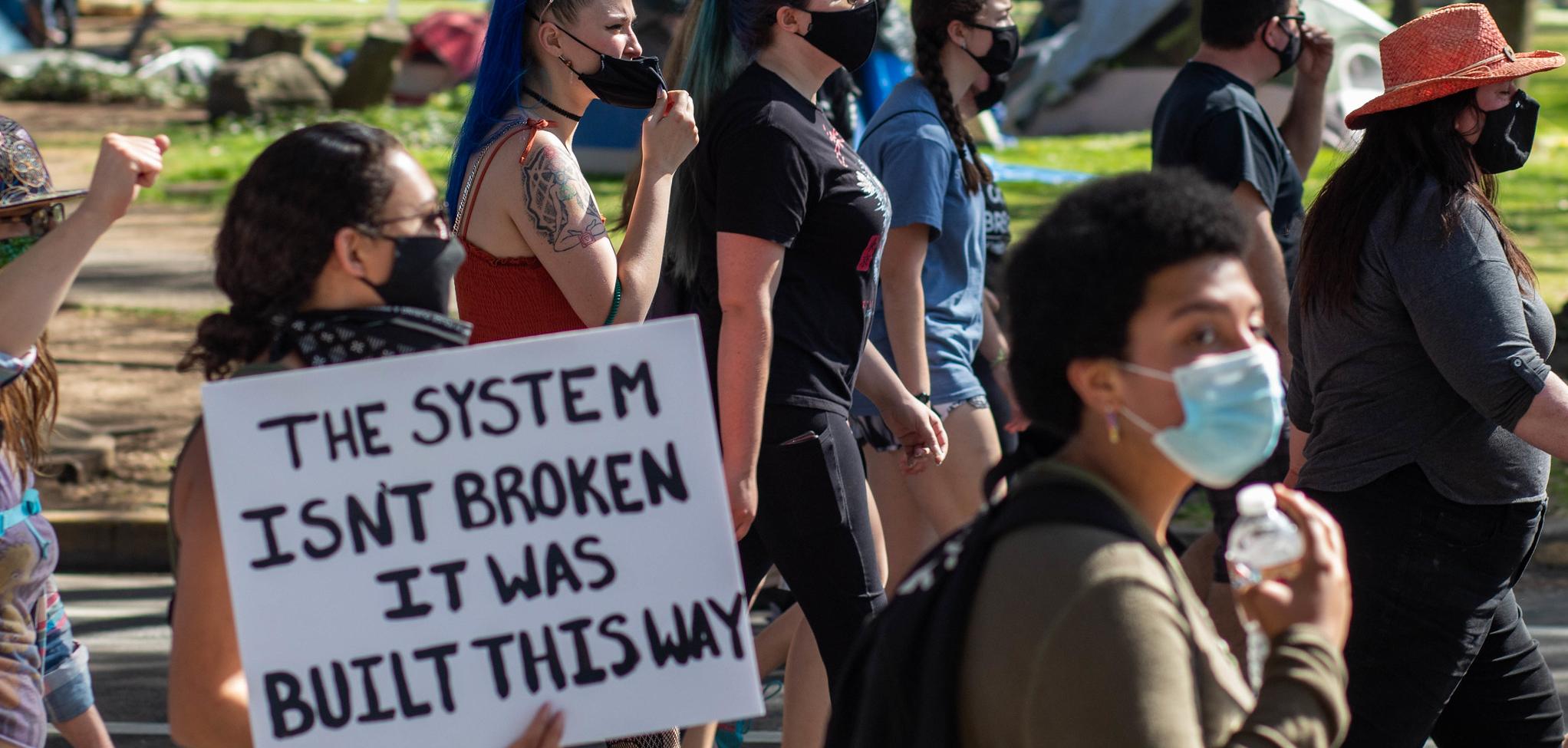 Protesters wearing masks. One protester holding a sign that says "The system isn't broken it was built this way."