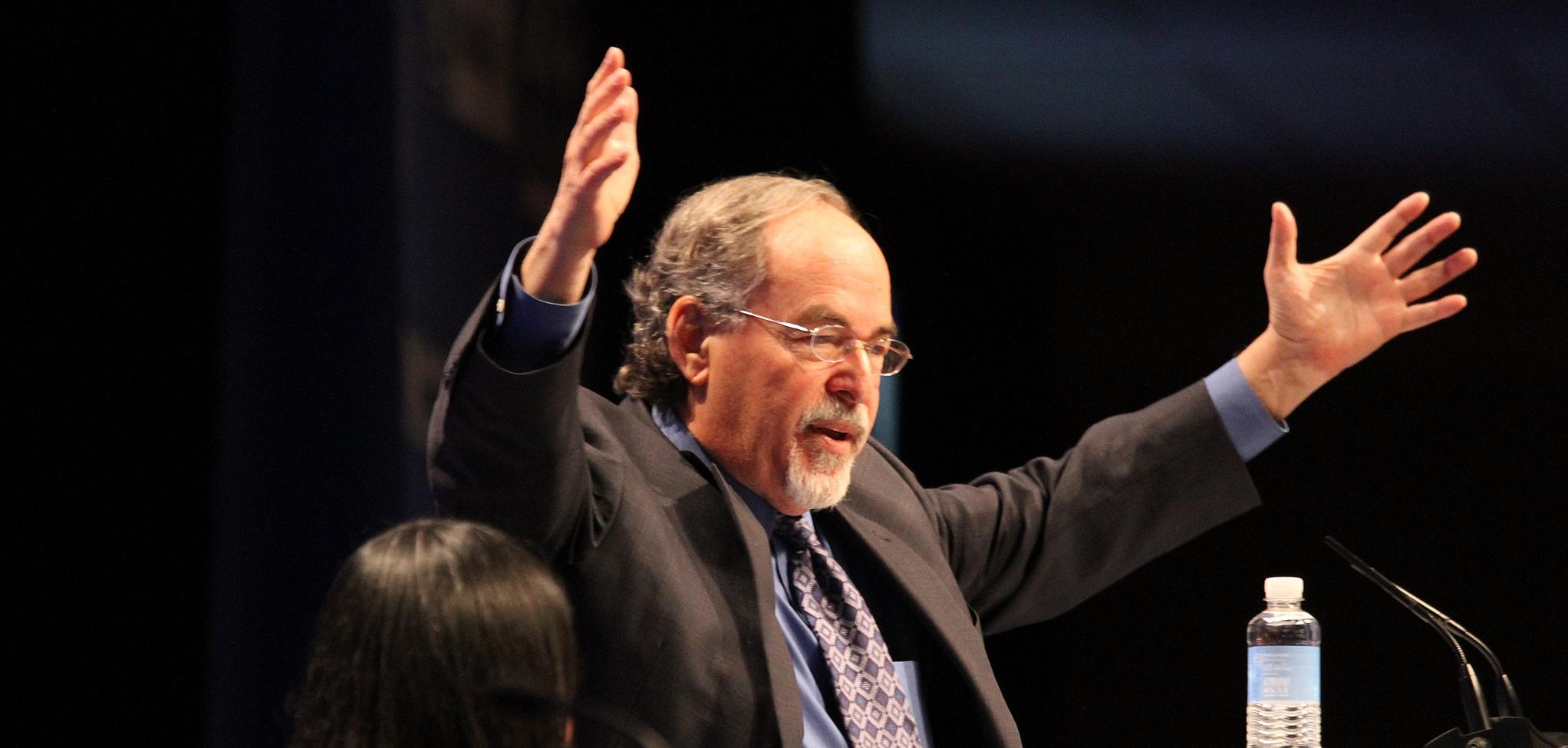 David Horowitz, a white man with his hands raised in the air, in front of a mic stand.