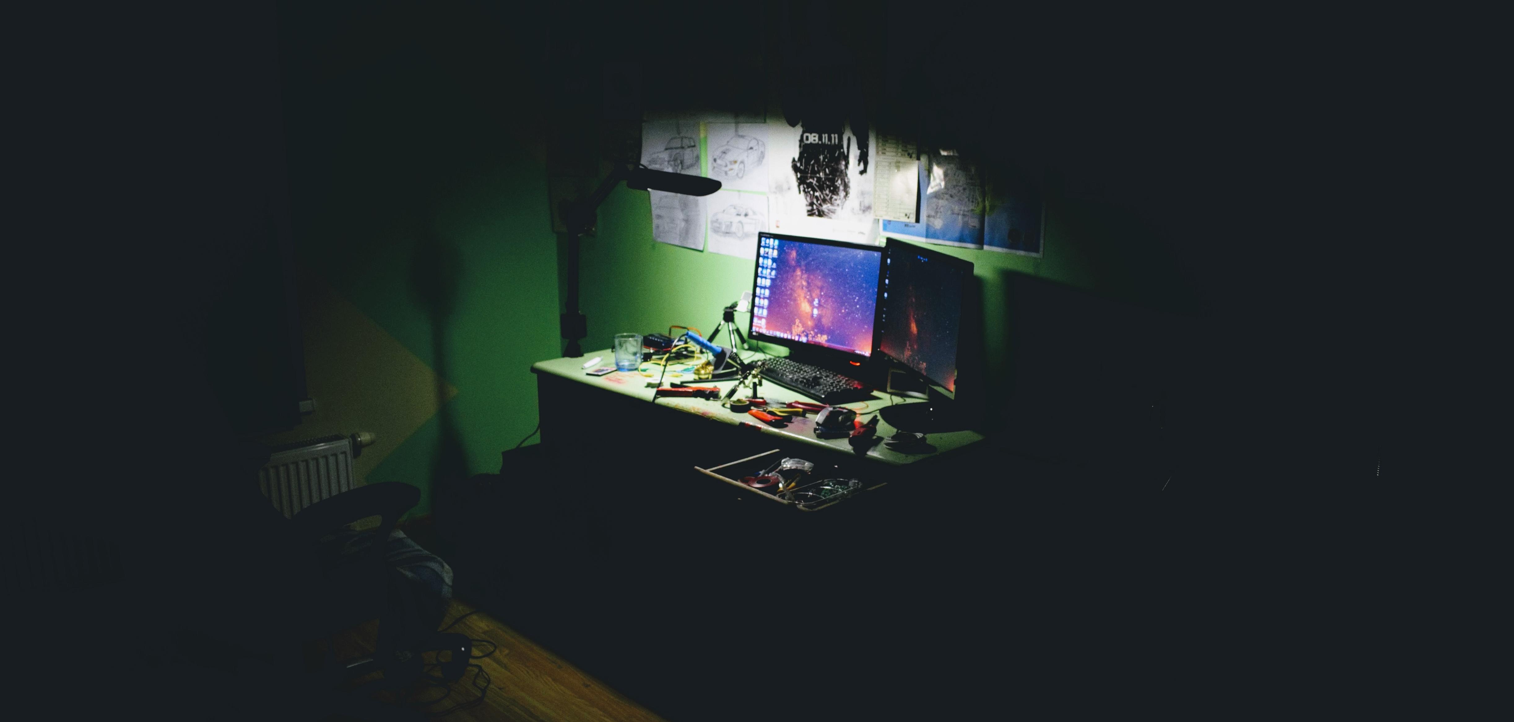 Two computer screens on a desk in a dark room. The light is focused on the computers